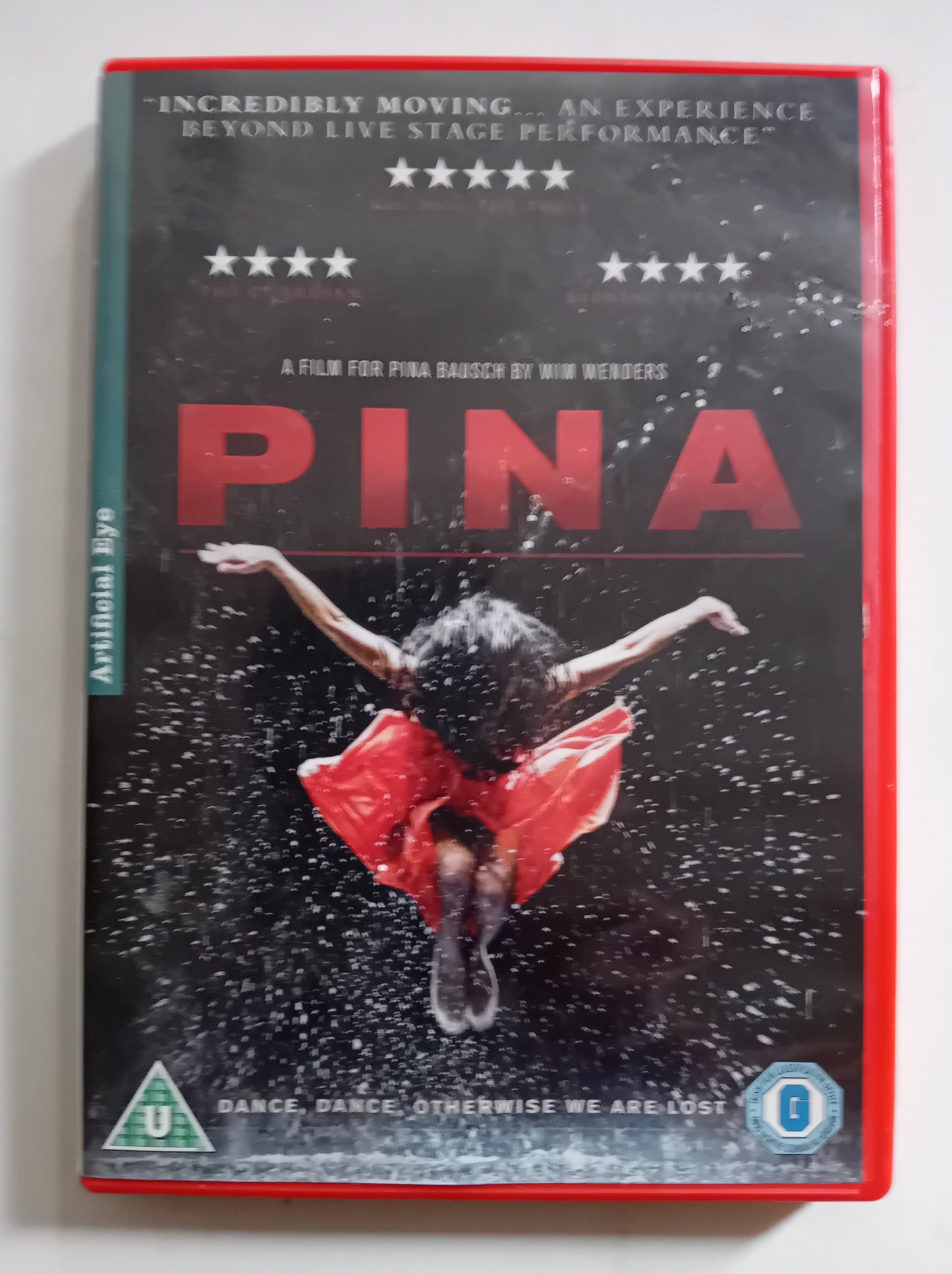 A DVD case for the film 'Pina' lying flat on a white surface. The cover is predominantly black with a dancer in a red dress mid-leap, large red title text, and critical review quotes at the top.