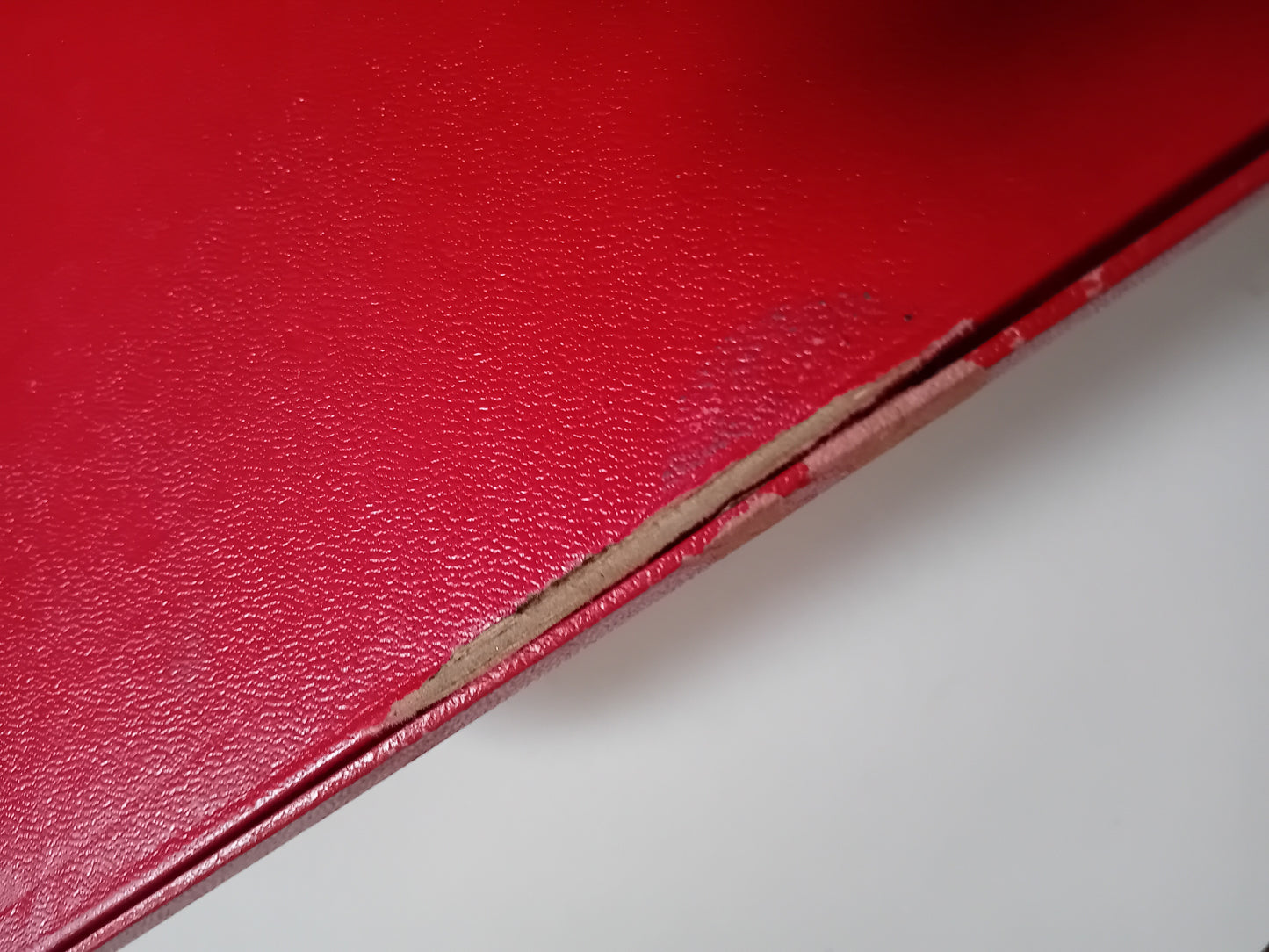 Close-up of a red textured leather or faux-leather surface with a visible edge and a small amount of worn, tan material along the edge, set against a light gray background.