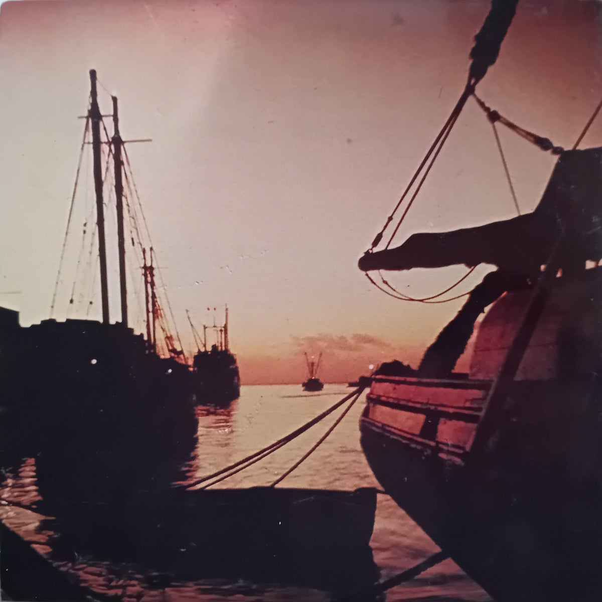 A vintage photograph of a harbor scene at sunset, featuring silhouetted boats and ships against a warm pinkish-orange sky, with the foreground showing part of a boat's hull and rigging.