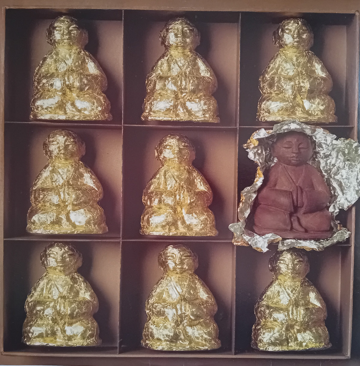 A boxed set of nine small seated Buddha figurines arranged in a 3x3 grid. Eight figurines are wrapped in shiny gold foil, while one in the bottom-right position is exposed and painted in a muted reddish-brown tone.