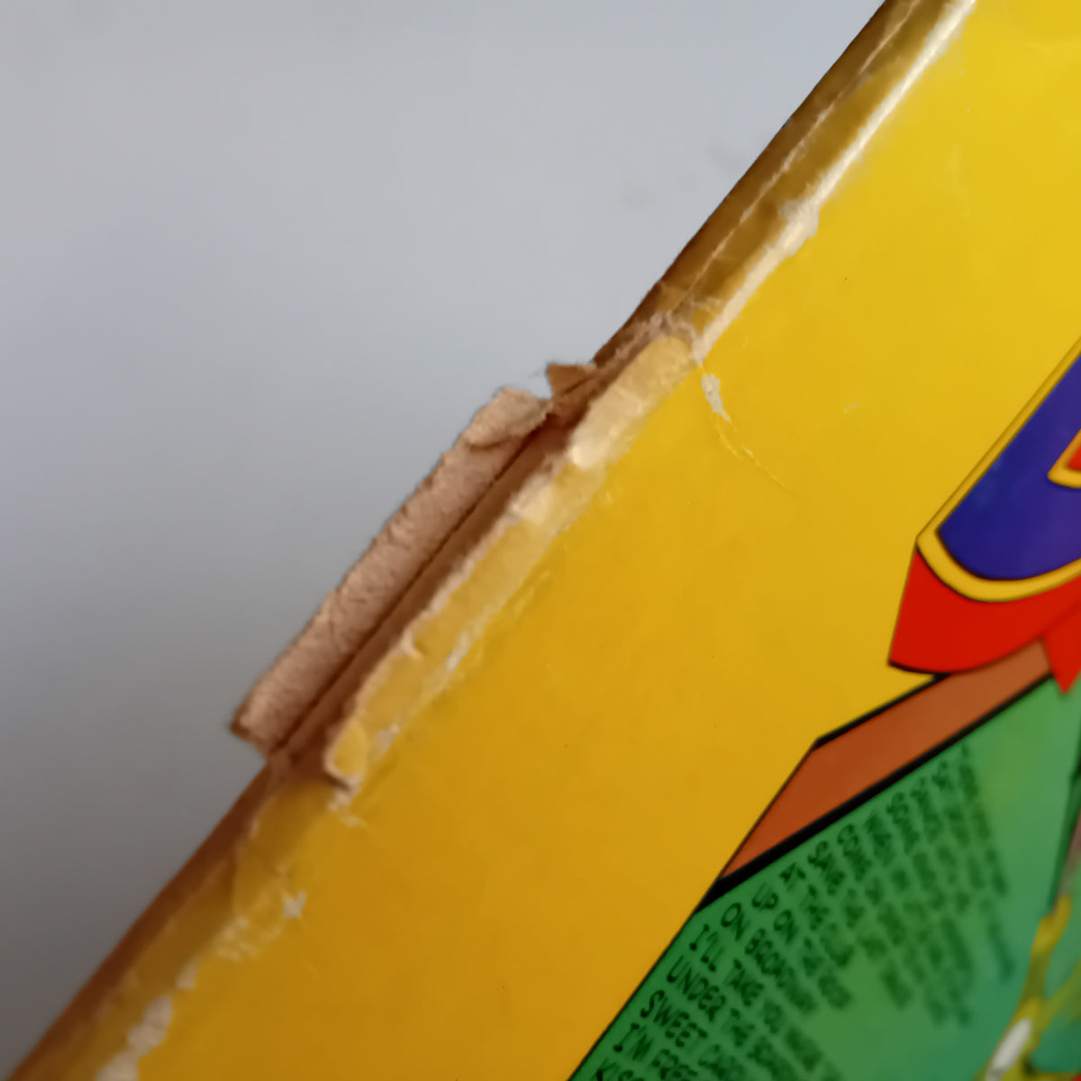 Close-up of a yellow cardboard sleeve spine with visible wear, scratches, and a torn edge, against a light gray background. Part of a colourful printed design is visible.