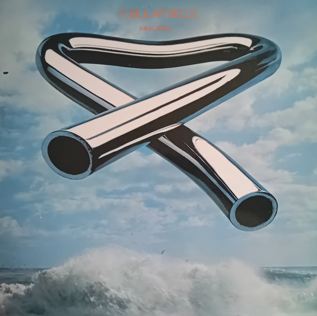 Album cover for Mike Oldfield's 'Tubular Bells' featuring a chrome metallic X-shaped object floating over an ocean scene with waves under a cloudy blue sky.