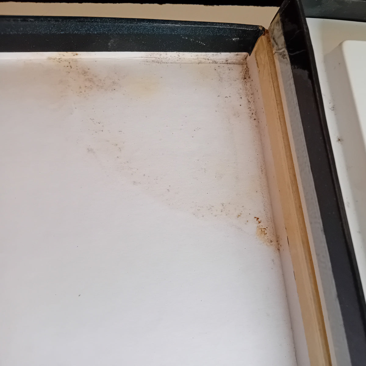 Close-up of the corner of a white box with a black trim and a light wood edge, showing brown dust, grime, and residue on the white surface.