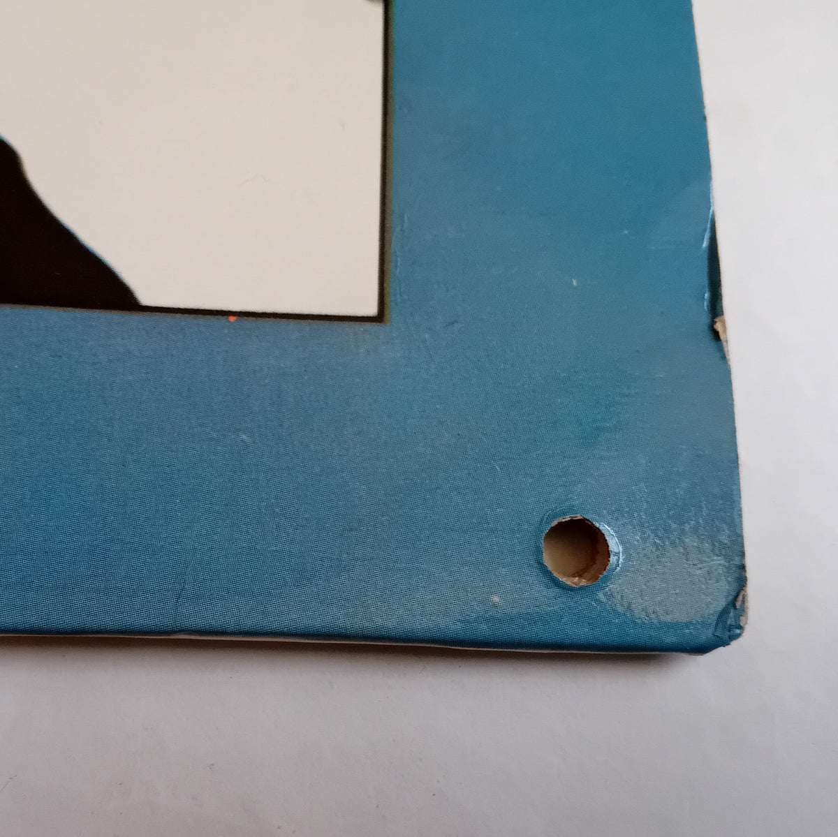 Close-up of the corner of a blue LP cover with a pre-drilled hole near the bottom right, on a light gray background.