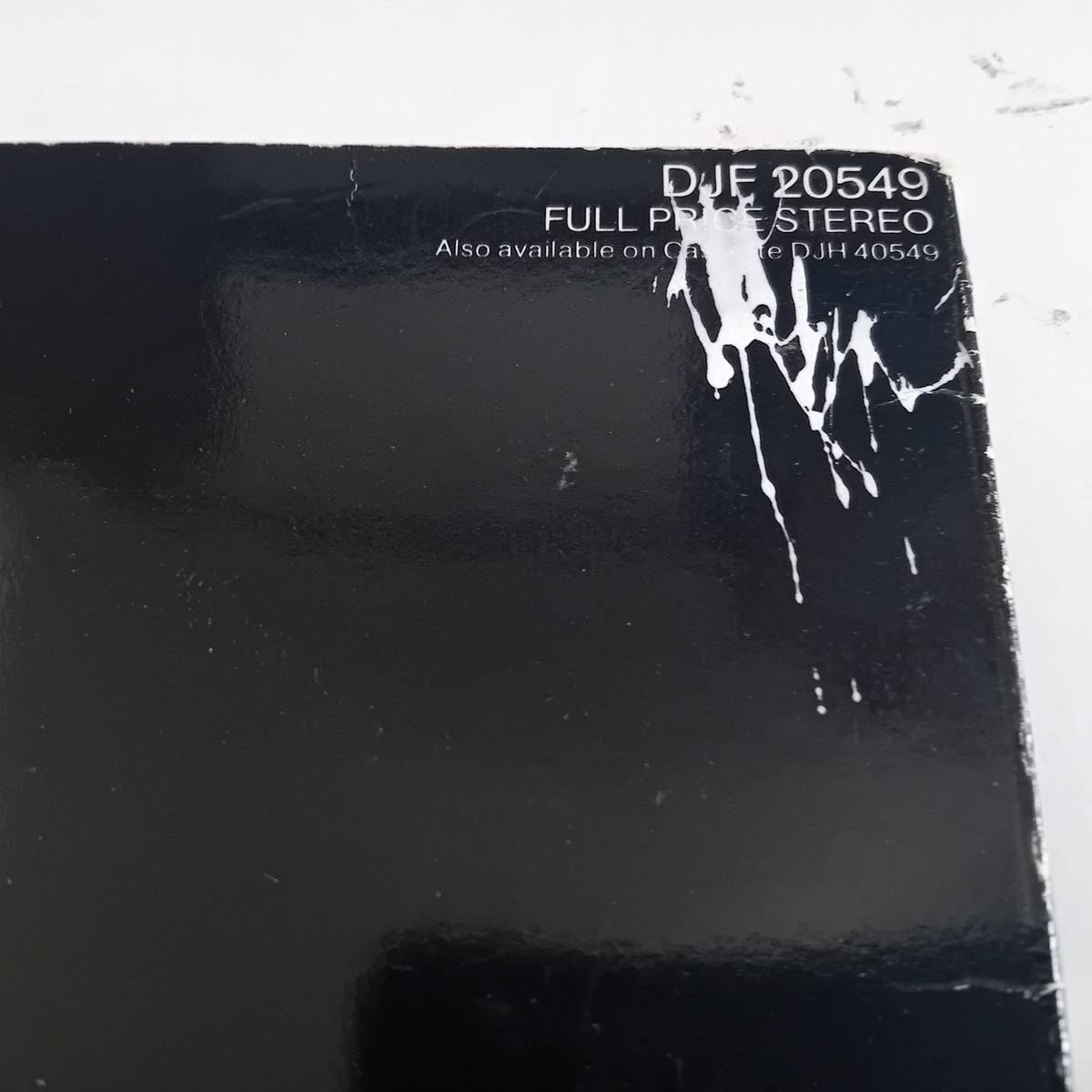 A close-up of a glossy black vinyl record sleeve corner with white printed text reading 'DJE 20549 FULL PRICE STEREO' and a white paint mark near the top right, photographed against a light off-white background.