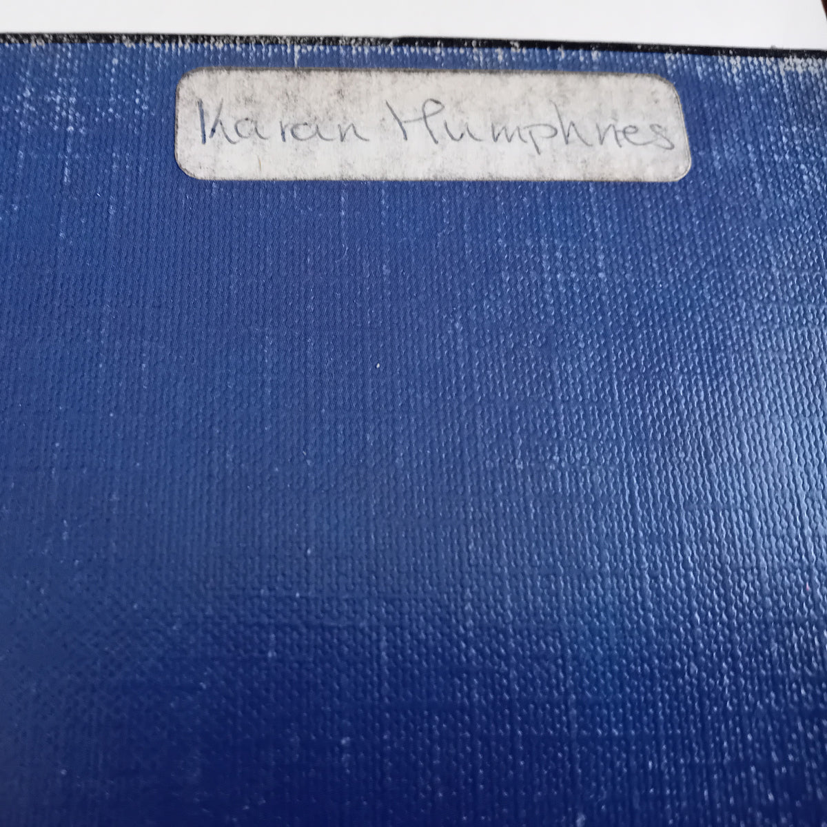A close-up of a blue textured fabric or canvas material with a white rectangular label near the top edge that reads 'Karen Humphries' in handwritten text.
