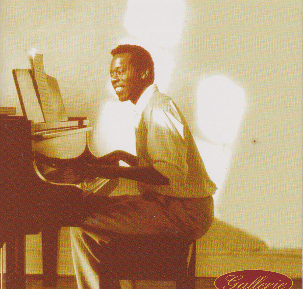 A sepia-toned photograph of a man seated at a grand piano, smiling and playing in a gallery setting. The image has a warm vintage aesthetic with strong light and shadow effects, and the word 'Galerie' appears in a red oval at the bottom right.