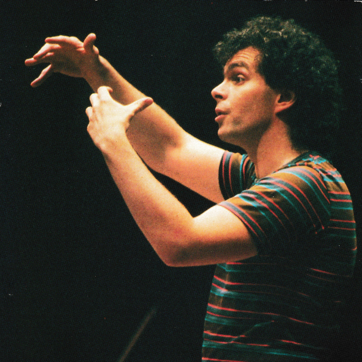 A man with curly dark hair wearing a green, orange, and blue striped t-shirt, gesturing with his hands against a dark background.