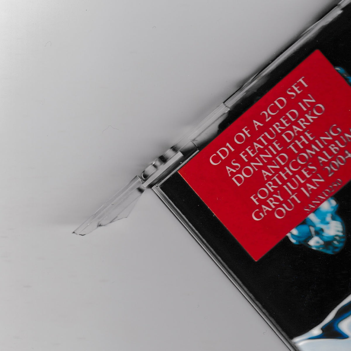 Close-up of a CD case corner with a red label reading 'CD1 OF A 2CD SET AS FEATURED IN DONNIE DARKO AND THE FORTHCOMING MEMORABLES' on a light gray background.
