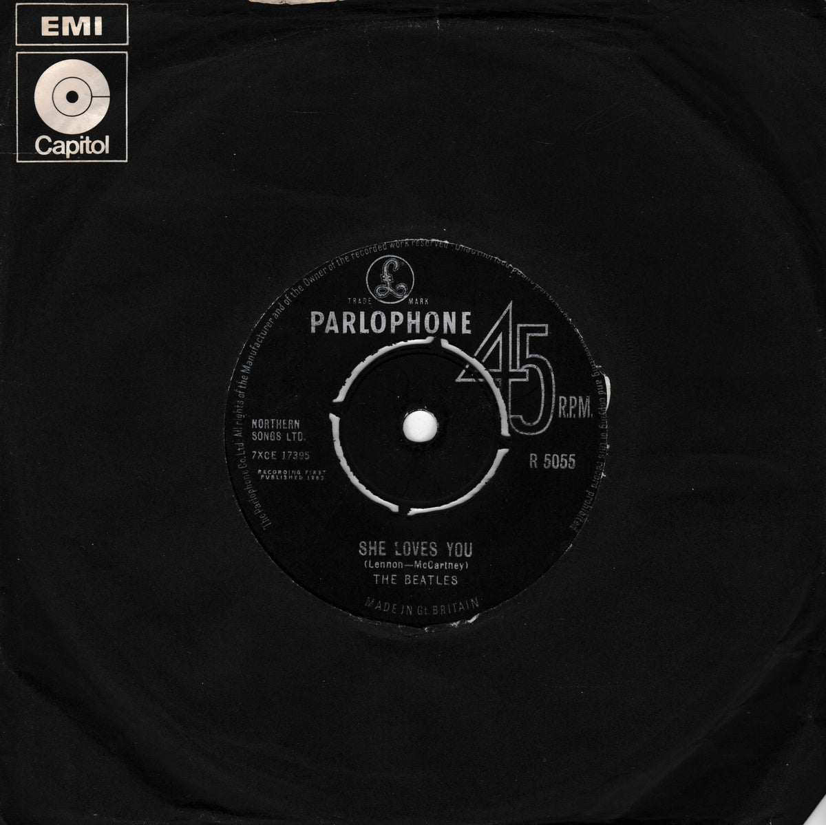 A black vinyl record partially pulled from its black paper sleeve, showing the Capitol Parlophone label with the Beatles' song 'She Loves You' and a Capitol EMI logo in the top-left corner.